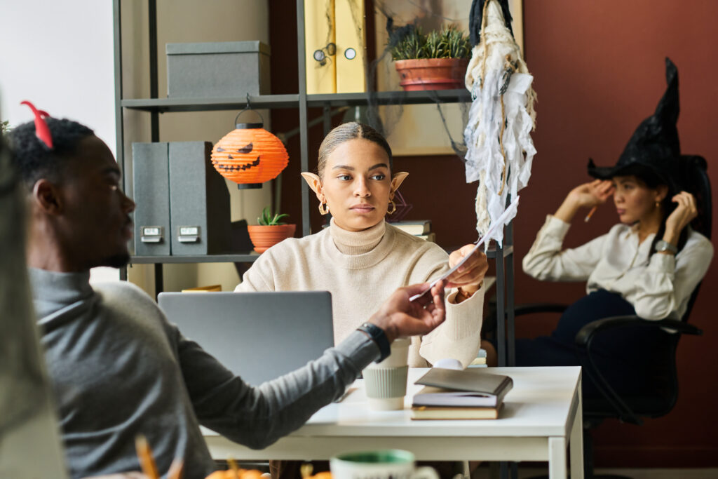 Three coworkers in an office setting dressed in Halloween costumes. The woman in the center is wearing elf ears and is handing a document to a male colleague wearing devil horns on his head. In the background, another woman in a witch hat adjusts her headpiece. Halloween decorations, including a pumpkin lantern and cobwebs, add a festive atmosphere to the office