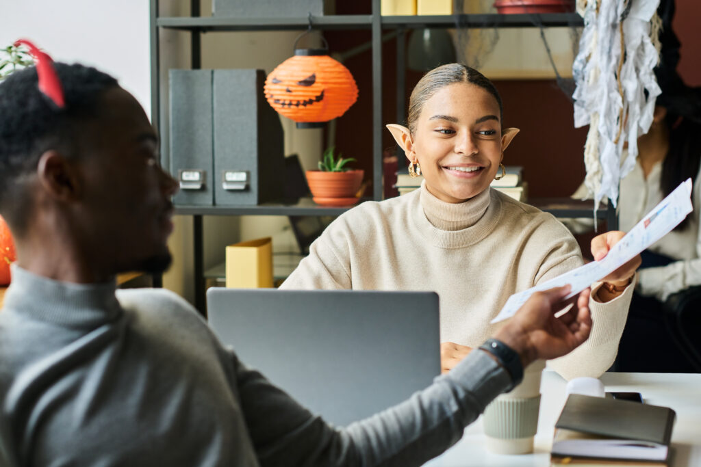 Office employees dressed in Halloween costumes working at their desks. A woman in the center wears elf ears and passes a document to a man on the left wearing devil horns. In the background, a woman in a witch hat adjusts her costume. Halloween decorations, such as a pumpkin lantern and cobwebs, add a festive atmosphere to the workspace.